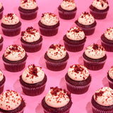 Row of Crumbs & Doilies Red Velvet Cupcakes with cream cheese frosting and red velvet crumbs on pink background